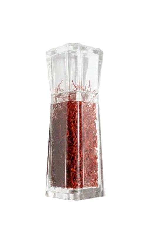 A coffin-shaped clear plastic container filled with red glitter or resin, photographed on a gray tiled floor with sunlight and shadows. A hand holding a patterned object is partially visible on the left edge.