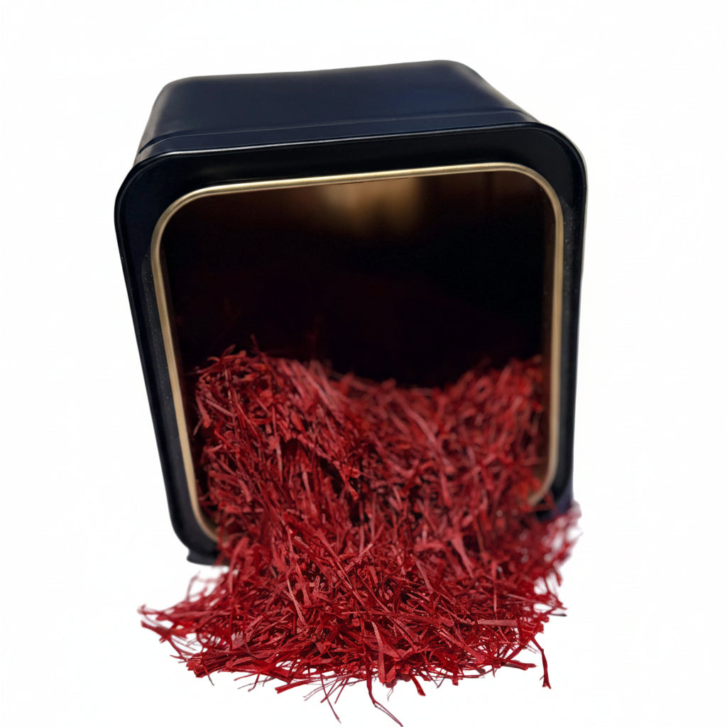 A small black square tin with a gold-toned rim, tipped over so that red saffron threads spill out onto a white surface.